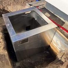 Ein quadratischer, metallischer Schacht ist in einem Erdloch installiert, umgeben von frischem Erdreich und Bauwerkzeugen.