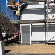 Ein graues Gehäuse steht an einer Baustelle, umgeben von Gerüst und unbefestigtem Boden unter strahlend blauem Himmel.