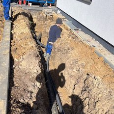 Ein Arbeiter steht in einem tiefen Graben, umgeben von Erde und Rohren, während er an einem Bauprojekt arbeitet.