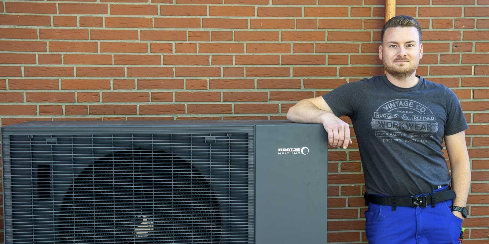 Vor einer Ziegelmauer posiert ein Mann in einem grauen T-Shirt und blauen Arbeitshosen neben einer Heizungsanlage.