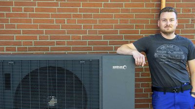 Vor einer Ziegelmauer posiert ein Mann in einem grauen T-Shirt und blauen Arbeitshosen neben einer Heizungsanlage.