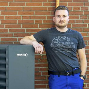 Vor einer Ziegelmauer posiert ein Mann in einem grauen T-Shirt und blauen Arbeitshosen neben einer Heizungsanlage.