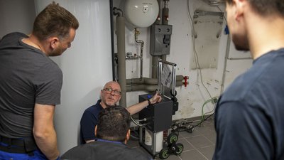 Eine Gruppe von Fachleuten diskutiert über eine technische Anlage in einem Keller, während einer von ihnen eine Anleitung zeigt.