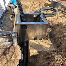 Ein rechteckiger, schwarzer Behälter steht in einem ausgehobenen Bereich, umgeben von Erde und Werkzeugen.