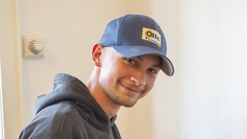 Der Mann trägt ein graues Sweatshirt und ein blaues Cap, sein Lächeln strahlt Freude aus.