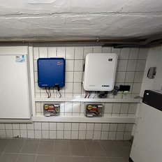 An einem weißen Fliesenwand sind zwei Wechselrichter montiert, einer blau und einer weiß, mit Kabeln und Zubehör darunter.