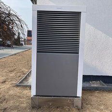 Ein moderner Wärmepumpen-Außenbereich mit einem grauen Gehäuse, auf einem gepflasterten Boden vor einem weißen Gebäude.