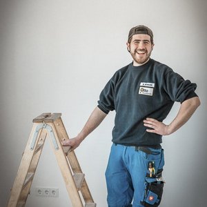 Ein Handwerker steht lächelnd neben einer Holzleiter, bereit für seine nächste Aufgabe in einem hellen Raum.