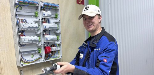 Ein junger Mann in blauer Arbeitskleidung lächelt, während er an einem elektrischen Schaltschrank arbeitet.