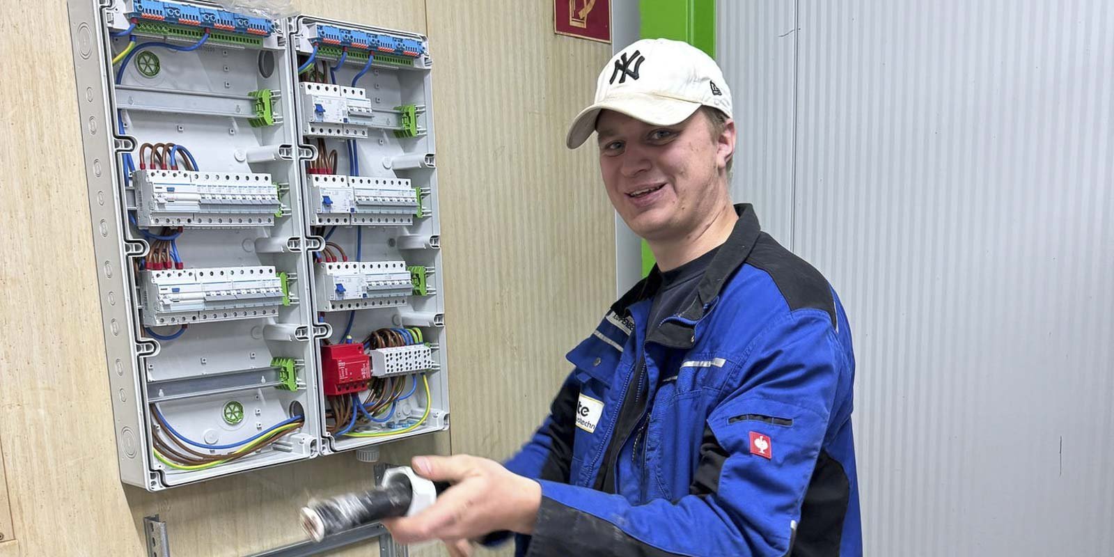 Ein junger Mann in blauer Arbeitskleidung lächelt, während er an einem elektrischen Schaltschrank arbeitet.