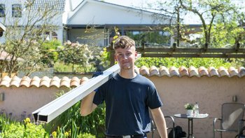 Ein junger Mann trägt ein langes Metallstück auf der Schulter und lächelt, während er in einem sonnigen Garten steht.