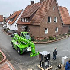 Ein grüner Kran steht auf einer ruhigen Straße vor einem Wohnhaus, während ein Mann in der Nähe steht und zuschaut.