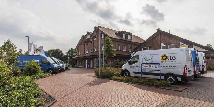 Ein modernes Gebäude mit roten Ziegeln, umgeben von grünen Pflanzen und mehreren Firmenfahrzeugen auf einem gepflasterten Parkplatz.