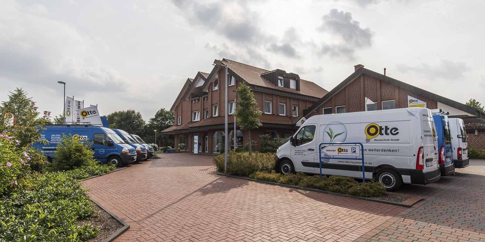 Ein modernes Gebäude mit roten Ziegeln, umgeben von grünen Pflanzen und mehreren Firmenfahrzeugen auf einem gepflasterten Parkplatz.