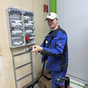 Ein Techniker in blauer Arbeitskleidung hält ein Werkzeug in der Hand und lächelt vor einem Schaltschrank mit Kabeln.