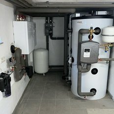 Ein Heizungsraum mit einem großen weißen Warmwasserspeicher und verschiedenen Rohren, die an die Wände angeschlossen sind.