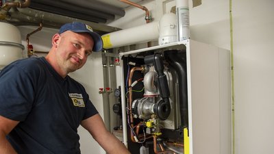 Ein Handwerker lächelt, während er an einer Heizungsanlage arbeitet, umgeben von Rohren und technischen Geräten.