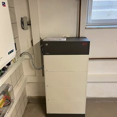 Ein moderner Batteriespeicher steht in einem hellen Raum, verbunden mit Kabeln und einem Stromanschluss.