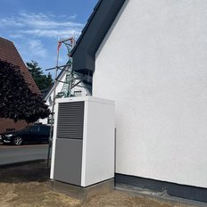 Eine moderne Wärmepumpe steht neben einem weißen Haus, während ein Gerüst im Hintergrund aufgestellt ist.