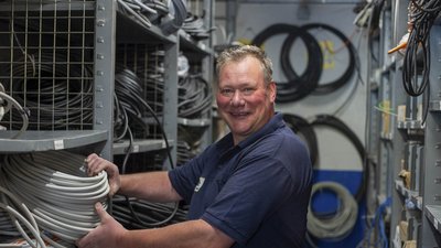 Ein lächelnder Mann sortiert Kabel in einem gut organisierten Lagerraum mit Regalen voller Kabelrollen.