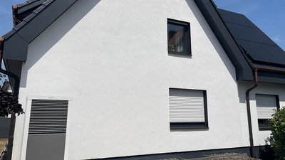 Ein modernes, weißes Haus mit einem schrägen Dach und Solarpanelen, umgeben von grünen Pflanzen und einem blauen Himmel.