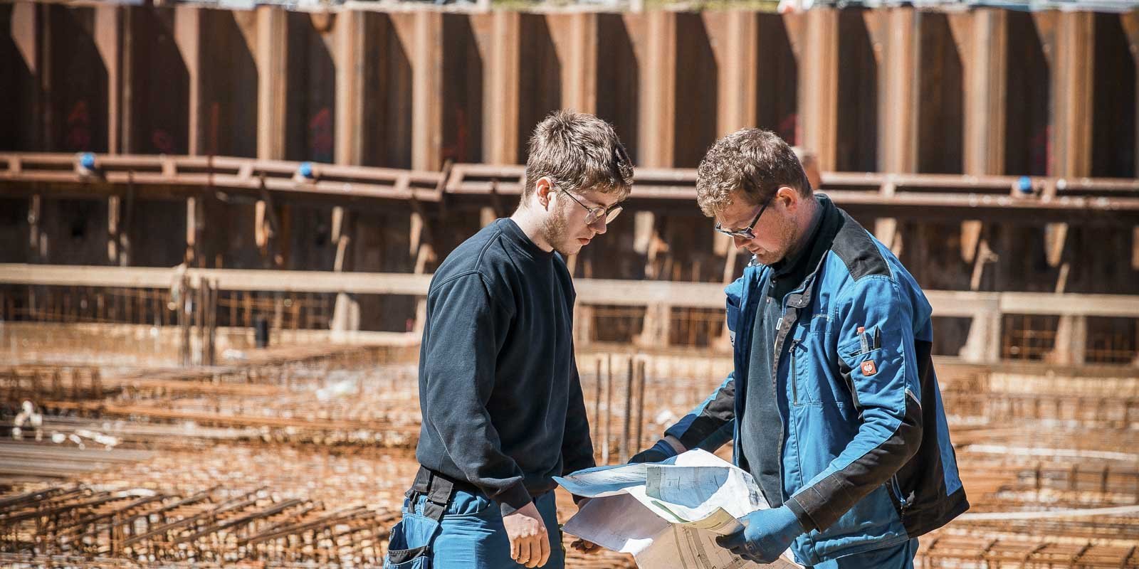 Zwei Bauarbeiter studieren Pläne auf einer Baustelle, umgeben von Stahlbewehrung und einem großen Baugrubenbereich.