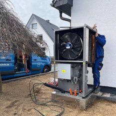 Ein Techniker installiert eine große Klimaanlage neben einem Wohnhaus, während ein Lieferwagen im Hintergrund parkt.