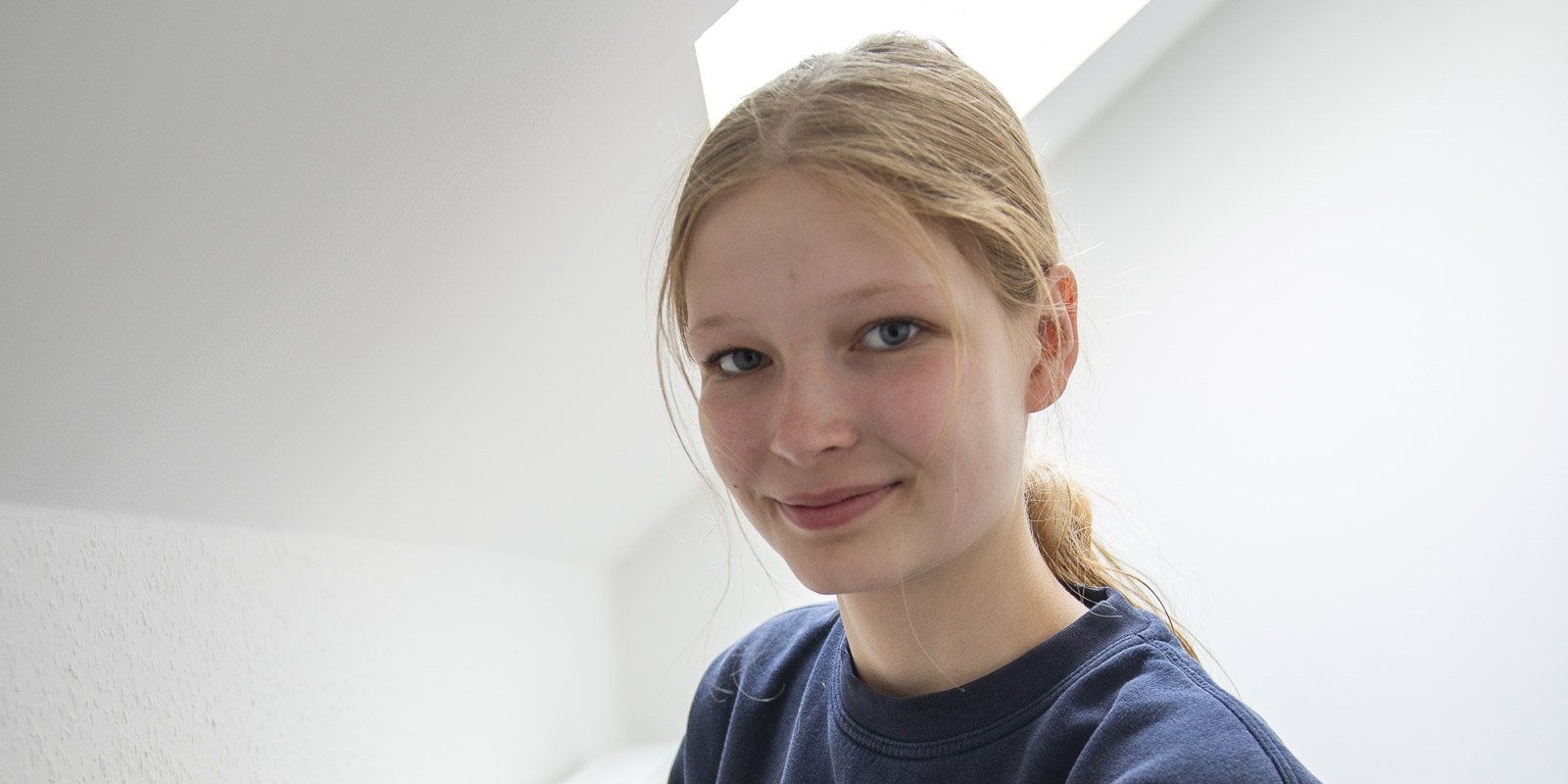 Ein lächelndes Mädchen mit blonden Haaren sitzt in einem hellen Raum mit schrägen Wänden und einem Fenster im Hintergrund.