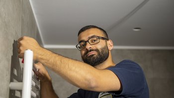 Ein Mann mit Brille montiert einen weißen Handtuchwärmer an einer grauen Wand in einem modernen Raum.