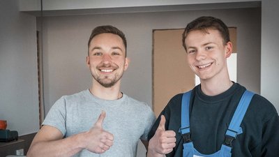 Zwei junge Männer stehen in einem Raum und zeigen Daumen hoch, lächelnd und zufrieden mit ihrer Arbeit.
