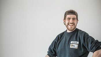 Ein junger Mann mit Bart und Baseballmütze lächelt freundlich, während er in einem hellen Raum steht.